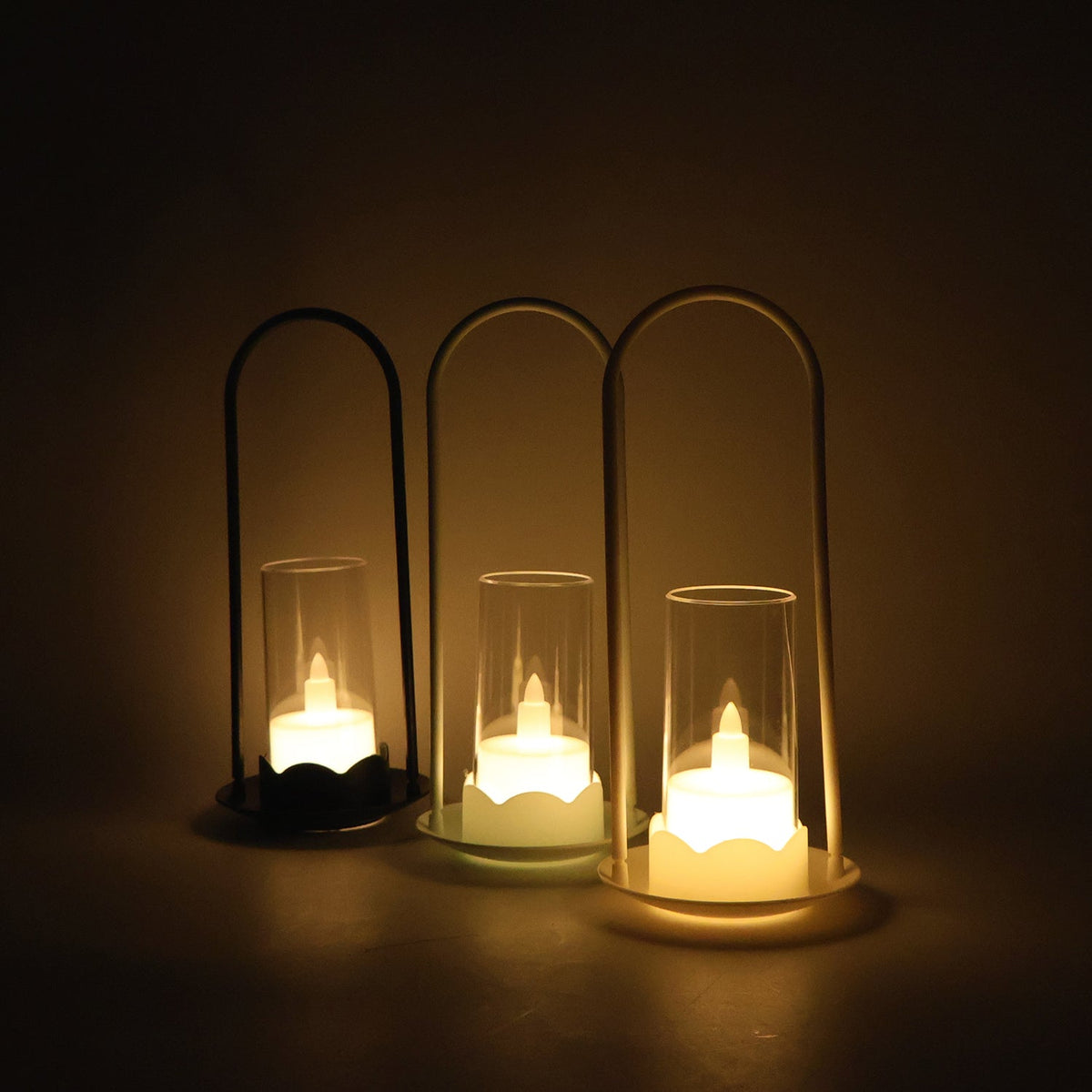 Three LED wind lanterns, one black and two white, with illuminated flameless candles, casting a warm glow in a dimly lit