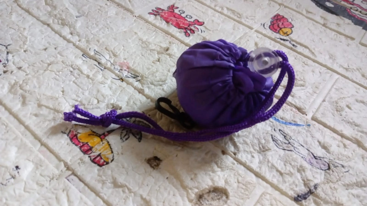 The purple foldable shopping bag is shown folded into a compact shape with a drawstring on a textured surface.