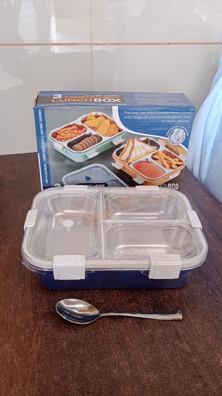 A lime green lunch box with three stainless steel compartments and dark clasps, with a spoon and fork, in front of its