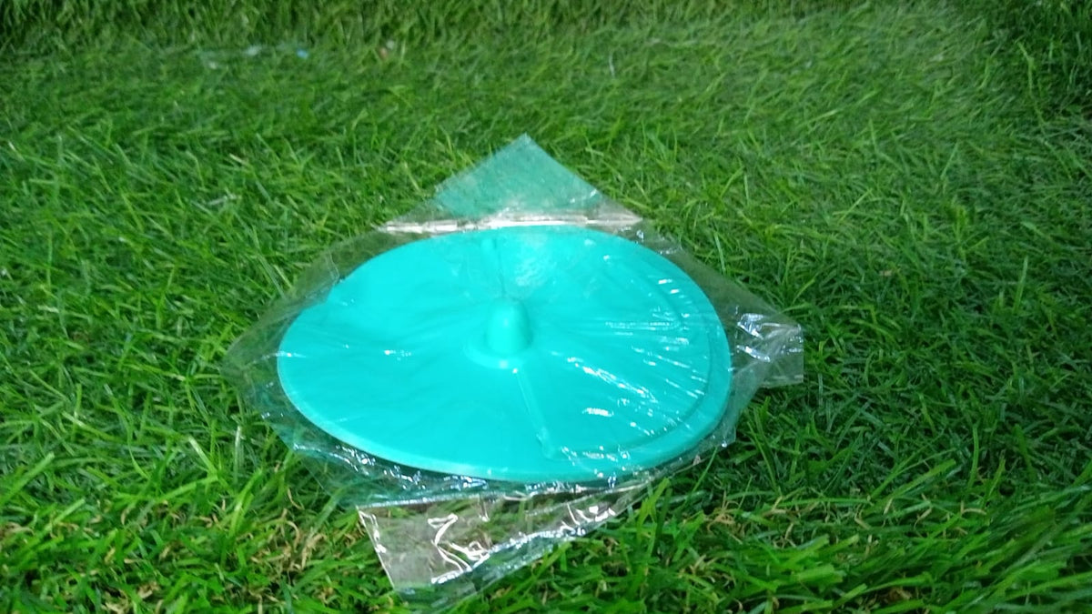 A teal silicone drain strainer, still in its clear plastic packaging, rests on vibrant green grass.