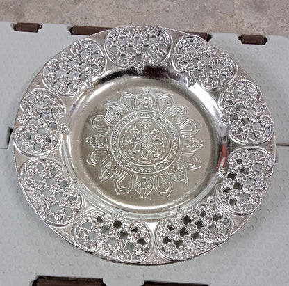 A silver-colored plastic pooja thali with a detailed embossed pattern resting on a light gray, textured surface.