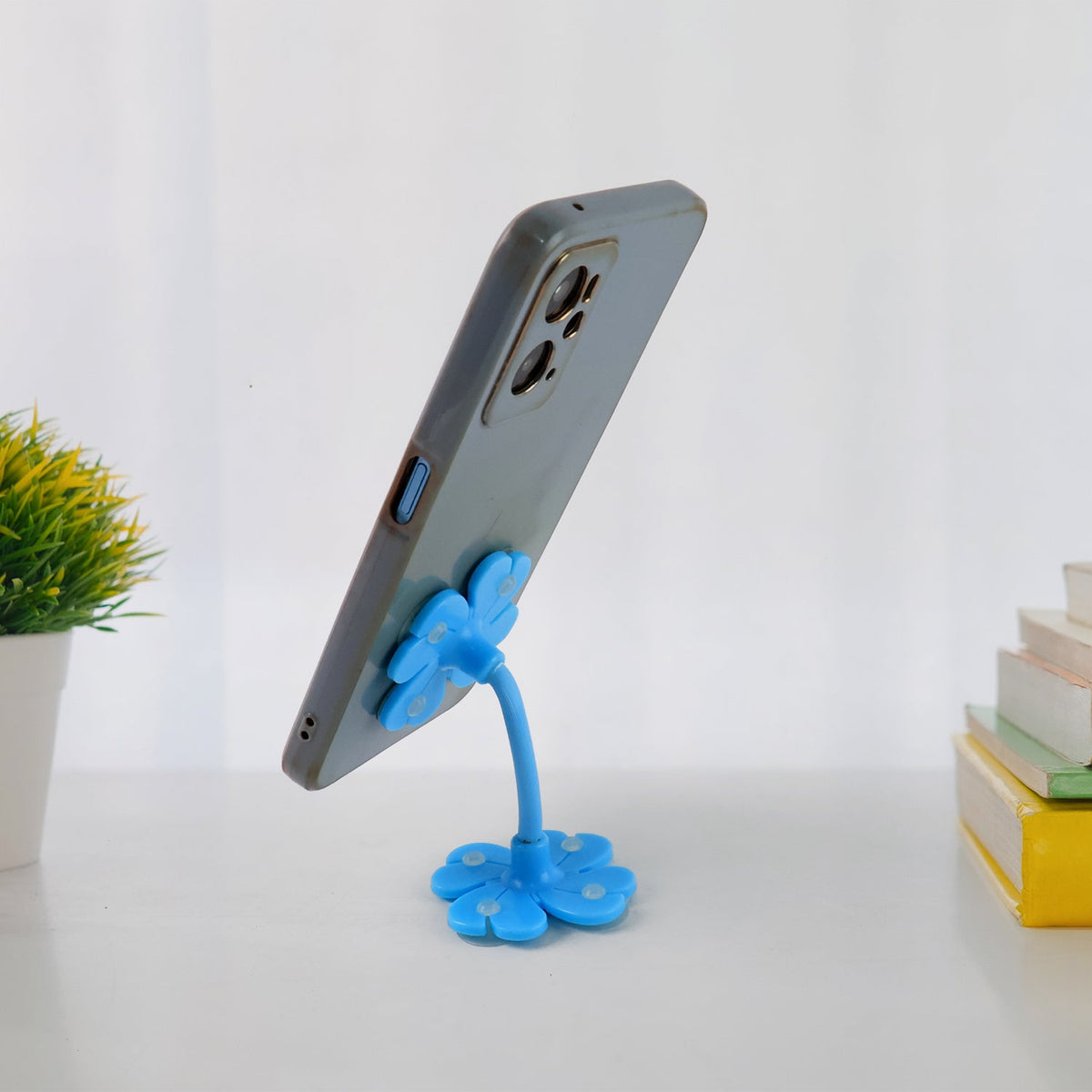 A gray phone is held by a blue flower-shaped phone holder with a flexible stem on a white surface with books.
