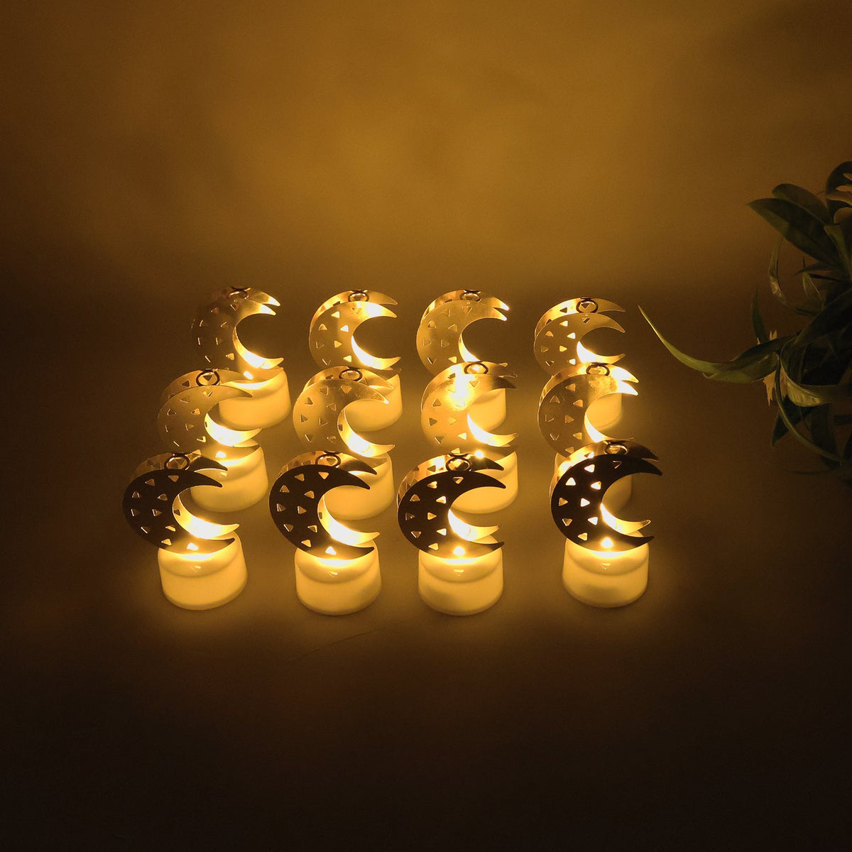 Ten gold moon shaped flameless LED candles with warm glow arranged on a dark surface, with a plant on the right.