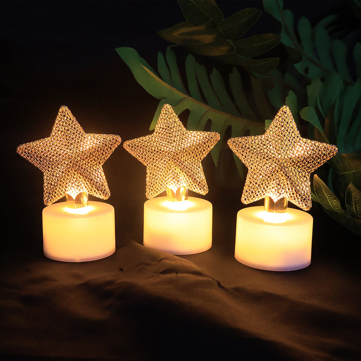 Three LED tealights with golden mesh stars glowing brightly against a dark green leafy background.