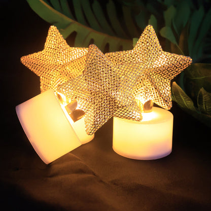 Three LED tealights with golden mesh stars, two standing and one tilted, glowing against dark foliage.