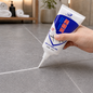 A hand applies white sealant from a tube to a gray tile floor grout line in a bathroom setting.