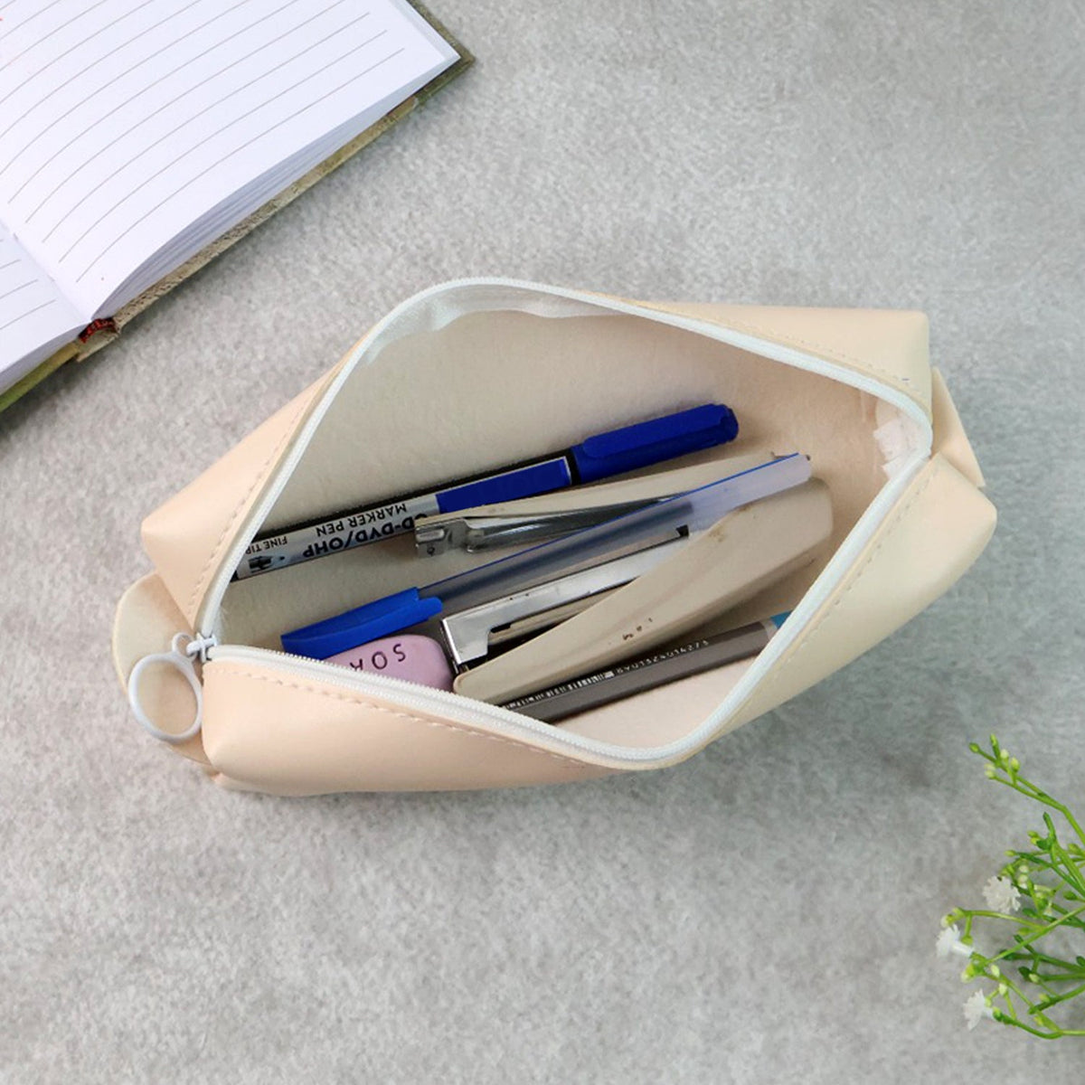 Open beige pencil case filled with pens, an eraser, and a stapler, next to a notebook.